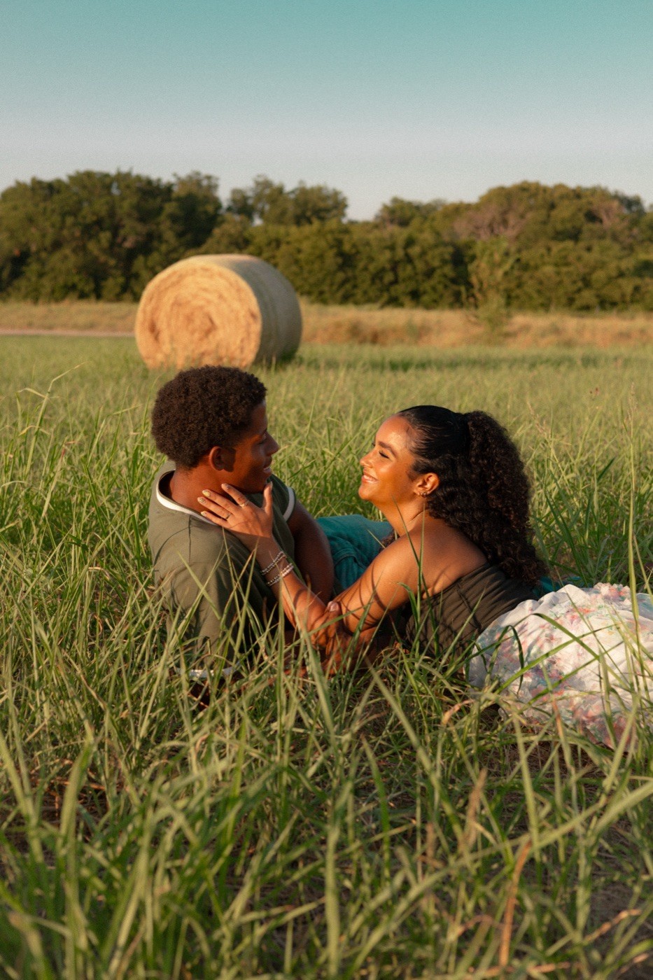 A couple lying together in tall grass beside a round hay bale.