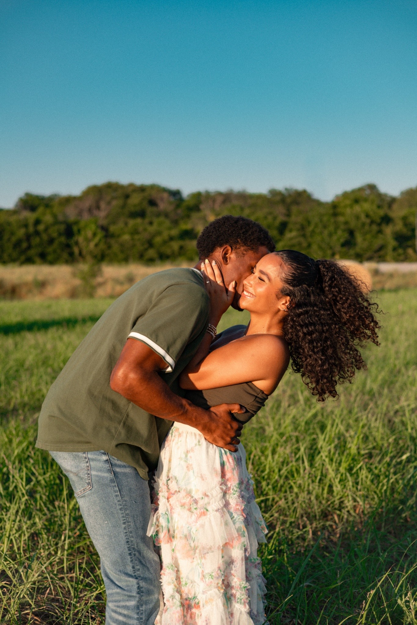 A couple kissing in a grass field under a clear blue sky.