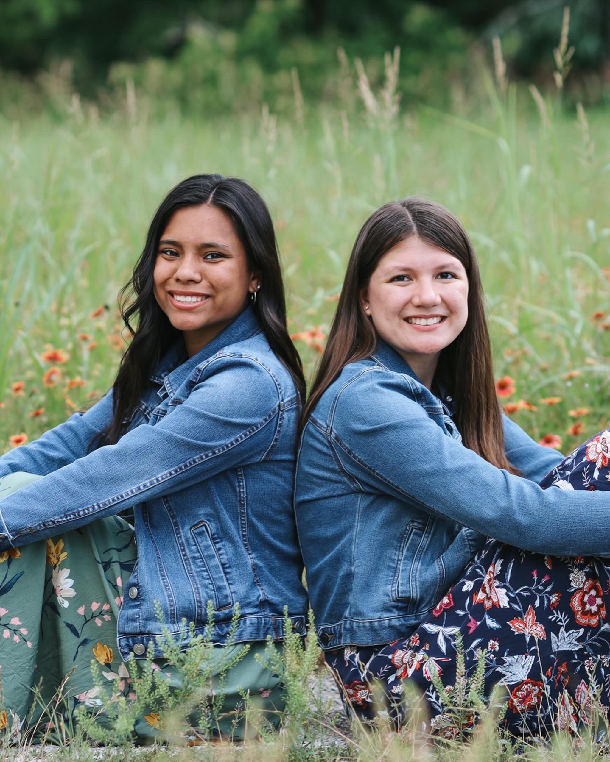 Two friends sitting back to back in a wildflower field, both in denim jackets.