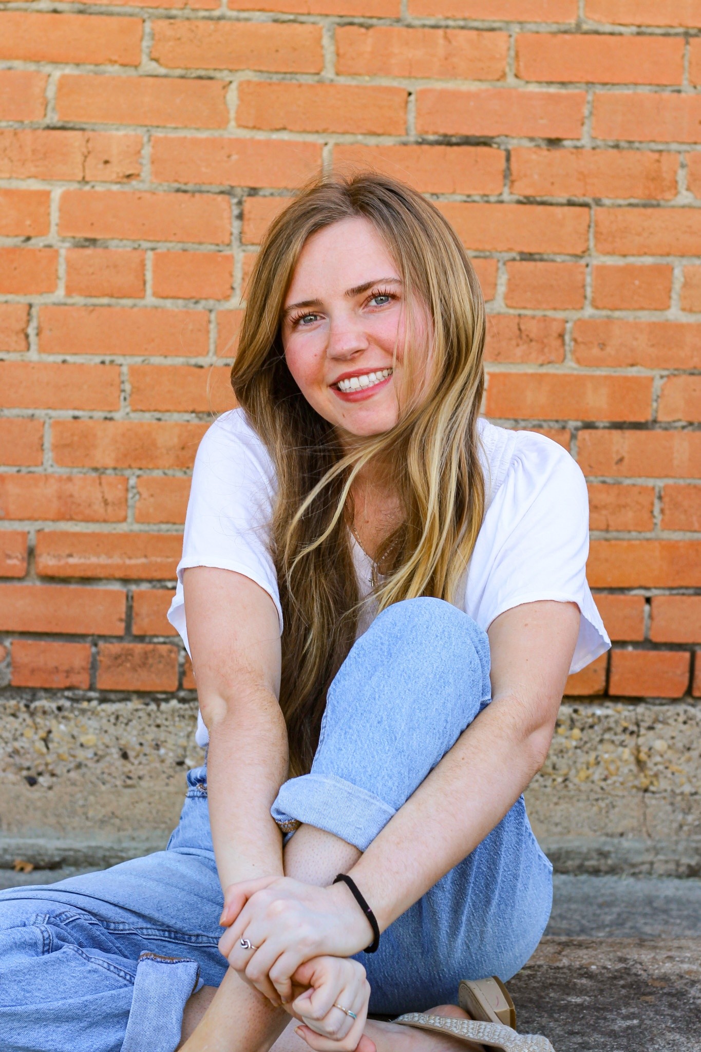 A woman smiling, seated against an orange brick wall.