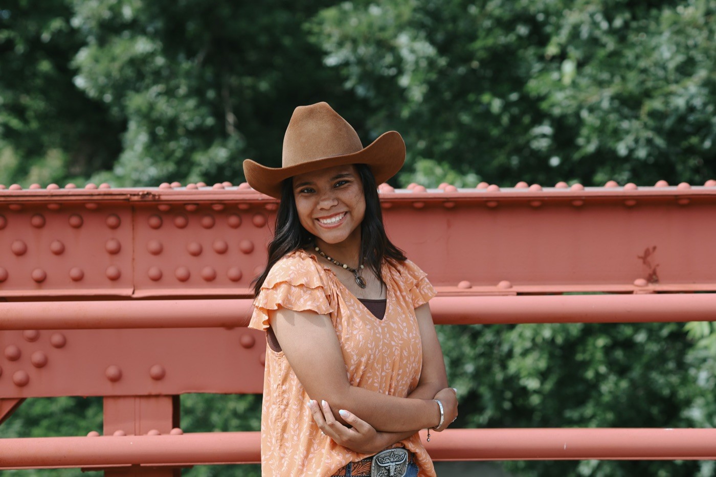 A senior in a cowboy hat leaning against a red steel bridge.