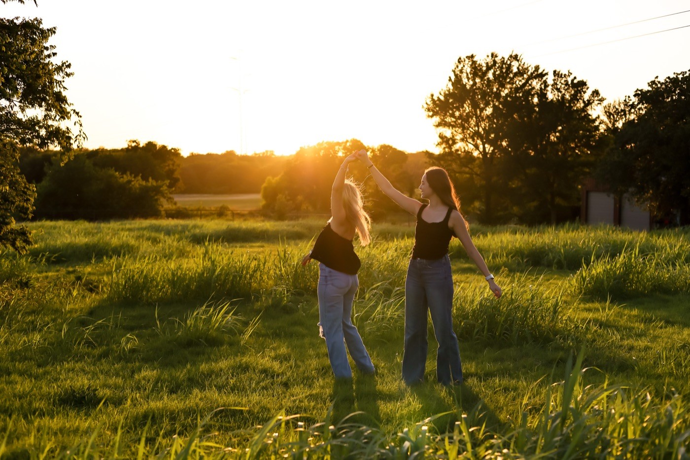 Two friends photographed small in a wide golden field as the sun drops behind the trees.
