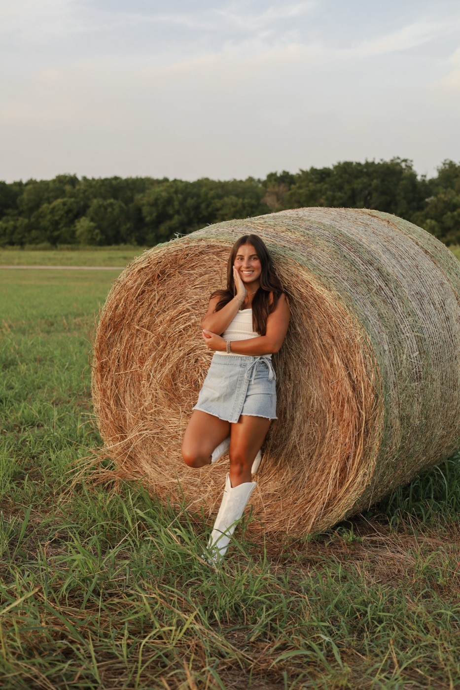 A senior leaning against a round hay bale in a mown field at dusk.
