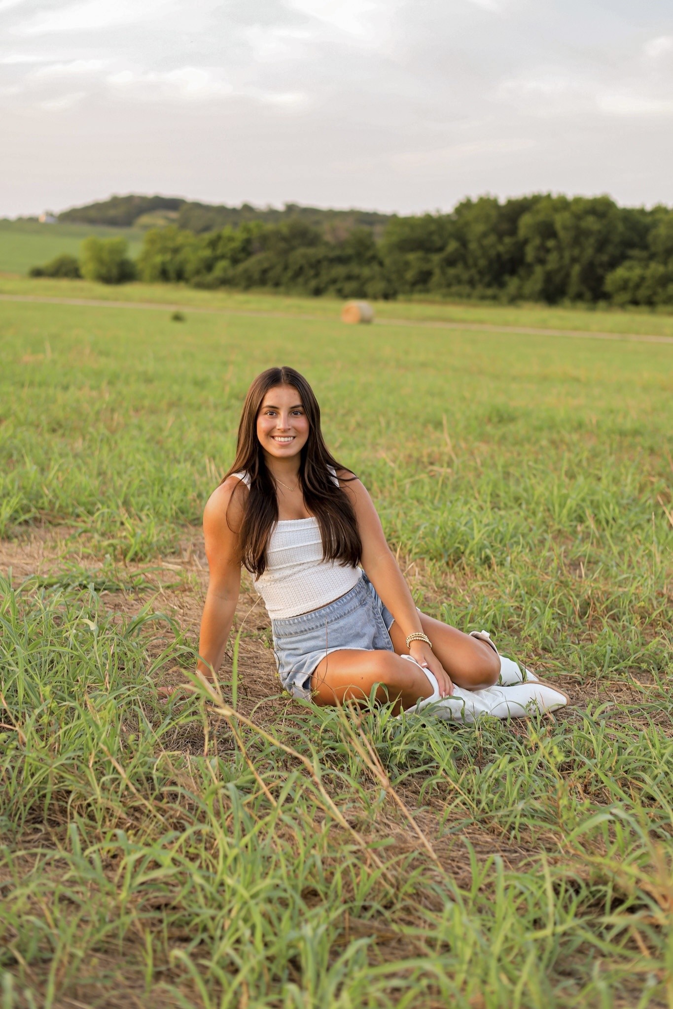 A senior sitting in tall grass with round hay bales in the distance at sunset.