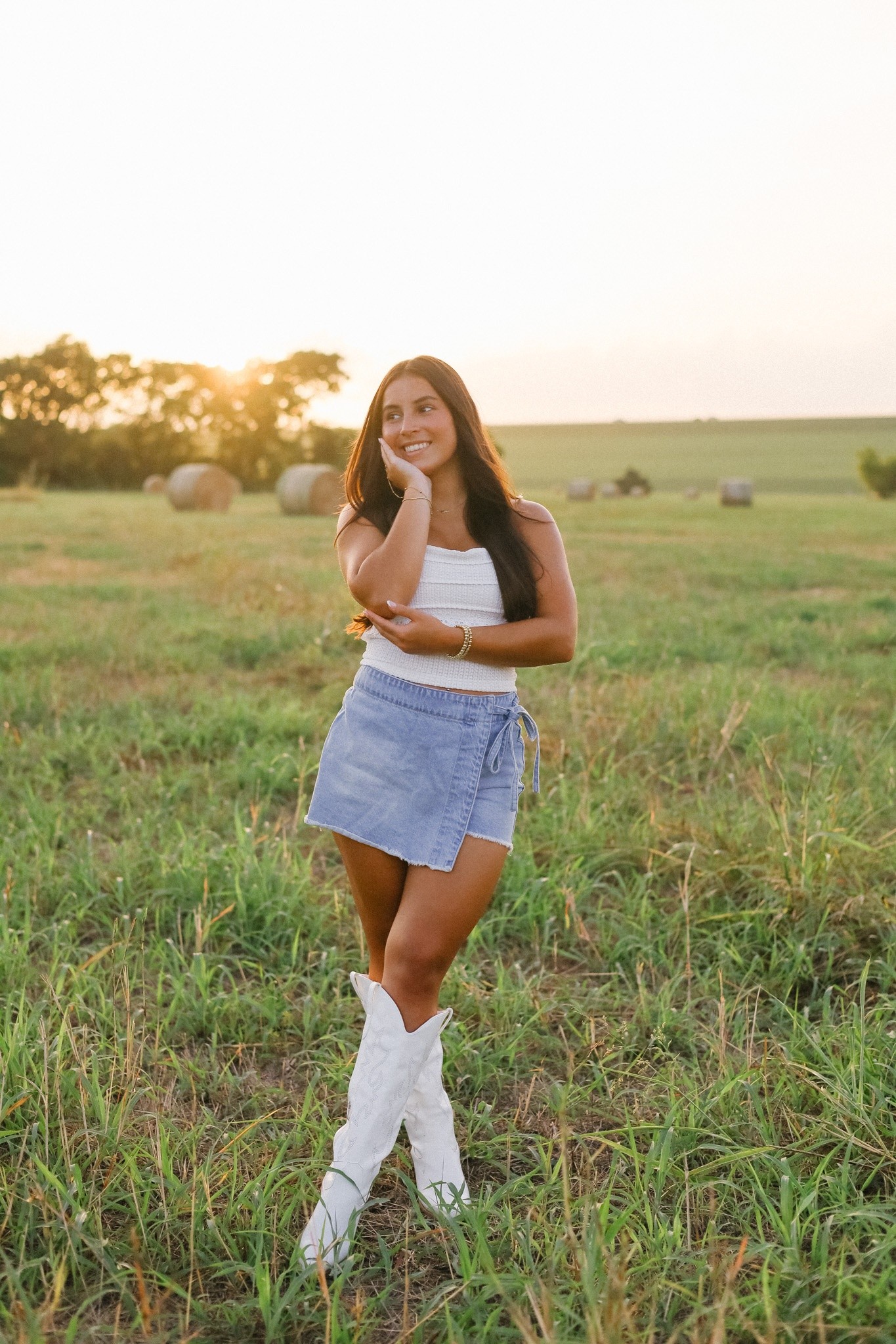 A senior in white boots and a denim mini skirt standing in a grass field at sunset.