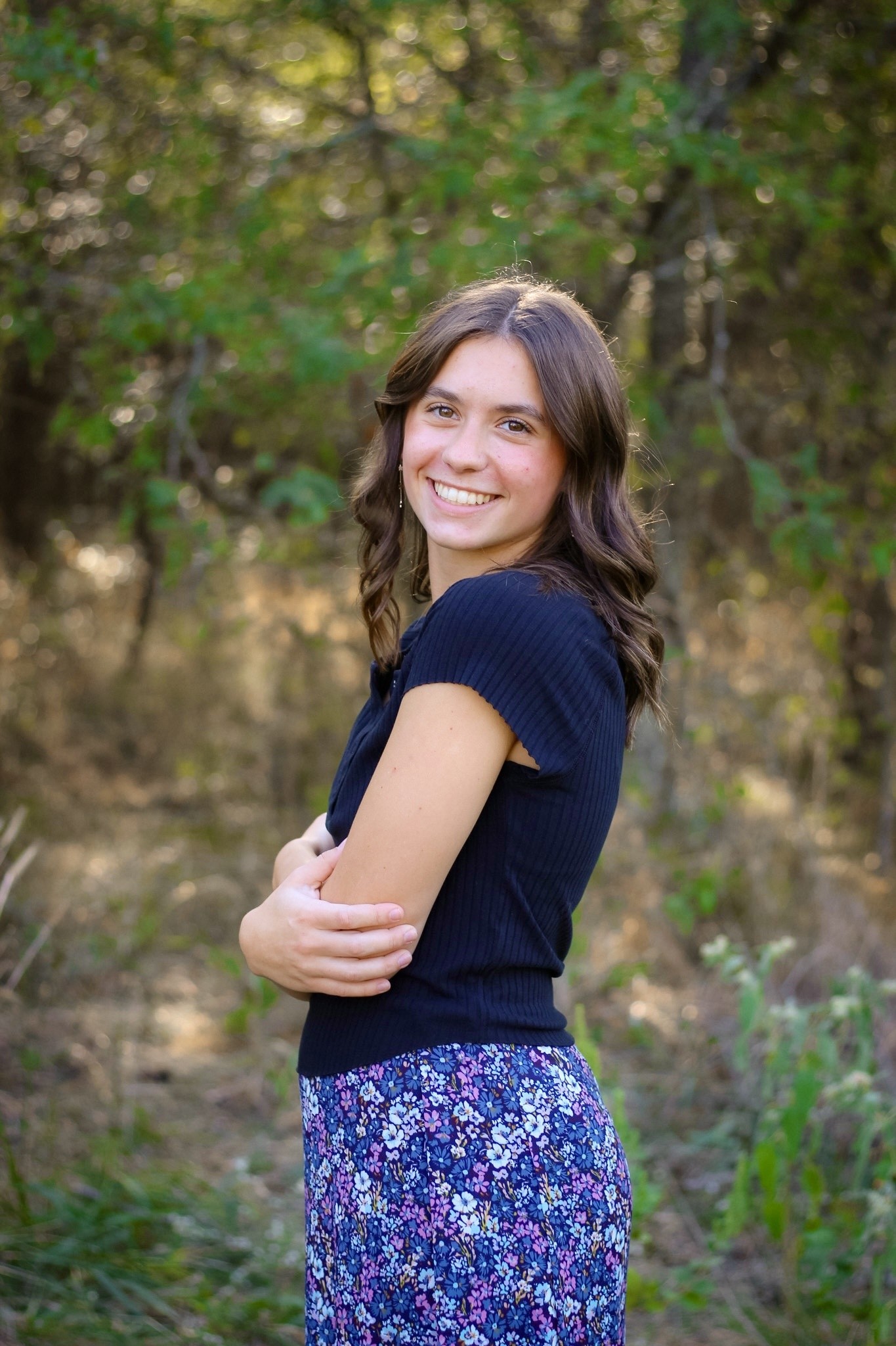 A senior with arms crossed, standing on a wooded path.