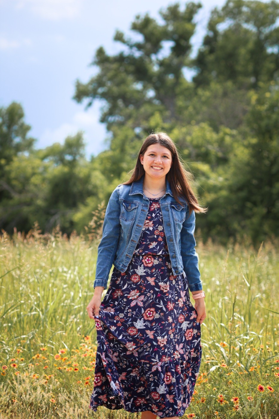 A young woman in a long floral dress standing among wildflowers on a summer evening.